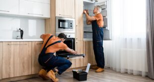 Safe and affordable repairs. Two handymen, workers in uniform fixing, installing furniture and equipment in the kitchen, using screwdriver indoors. Furniture repair and assembly concept